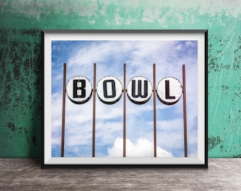 May include: A framed wall art print featuring four circular white signs mounted on thin brown metal poles against a blue sky with white clouds. Each sign displays a single black letter, collectively spelling the word BOWL in bold capital letters.