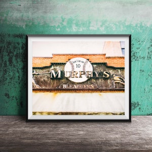Op de afbeelding: Een vintage, verweerd bord met de woorden "Murphy's Bleachers" in gouden letters. Een honkbal met het woord "believe" en het nummer "10" is gecentreerd op het bord.