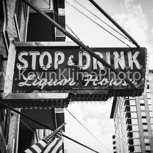 Op de afbeelding: Zwart-witfoto van een vintage neon-bord met de tekst "Stop & Drink Liquor House" dat aan een ketting boven een bakstenen gebouw hangt.