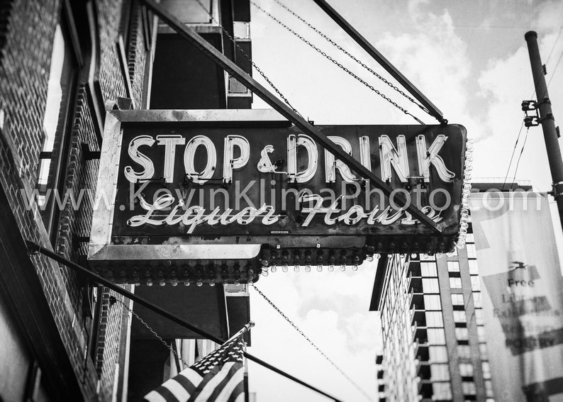 Op de afbeelding: Zwart-witfoto van een vintage neon-bord met de tekst "Stop & Drink Liquor House" dat aan een ketting boven een bakstenen gebouw hangt.