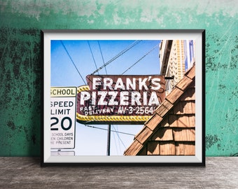 May include: A vintage sign for Frank's Pizzeria with a red and white background and yellow trim. The sign is hanging from a chain and is attached to a building with a wooden roof. A speed limit sign is visible in the foreground.