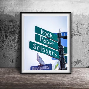 May include: Framed print featuring street signs with the words "Rock Paper Scissors" in white text on green backgrounds. Additional signs read "Decisions" and "Revisions". The artwork is displayed on a wooden surface.