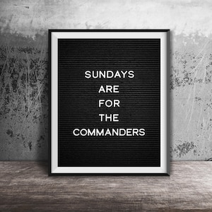 May include: A black letter board in a black frame with the white text "SUNDAYS ARE FOR THE COMMANDERS". The board has a white border. It is placed on a wooden surface against a grey concrete wall.