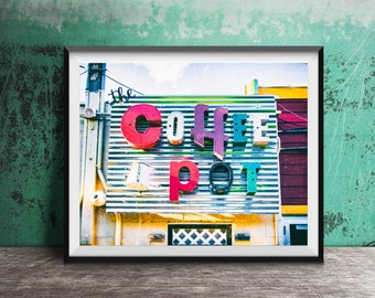 May include: A framed wall art print featuring a corrugated metal sign with the words "the COFFEE pot" in colorful, dimensional block letters. The sign includes a small white bucket graphic labeled "LUNCH" and is mounted on a building exterior.