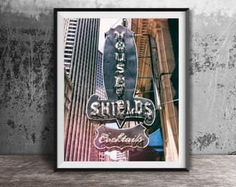 May include: A vintage-style sign for "House of Shields" Cocktails, with a black and white design and neon lights. The sign is hanging from a building in a city setting.
