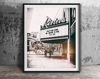 May include: A black and white framed print depicting the Riviera theater marquee with the text ONE YEAR DARK NO SHOWS SINCE 3 1 20, a large circular sculpture, and a Golden House Restaurant sign on a city street with two silhouetted figures walking.