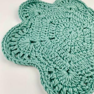 May include: A sage green crochet coaster featuring a flower-like shape with six rounded petals. The piece is crafted from thick yarn using a repeating stitch pattern that radiates from the centre, creating a textured, circular design.