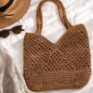 May include: A brown crocheted tote bag with an open-weave pattern, featuring two long shoulder straps. The bag is displayed alongside a straw hat with a black ribbon, brown tortoiseshell sunglasses, and a piece of light-colored fabric.