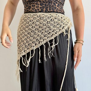 May include: A beige crochet hip scarf featuring an intricate star-like pattern with a triangular shape and long fringe along the bottom edge. The accessory includes a thin crocheted tie cord with a small wooden bead at the end for fastening.