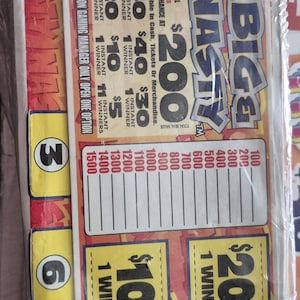 Puede incluir: Una hoja de boletos de juego Big Nasty envuelta en plástico transparente. El boleto presenta gráficos rojos y amarillos con texto que incluye Big Nasty, $200, $100 y varios montos de premios. Una etiqueta de código de barras blanca está arriba.