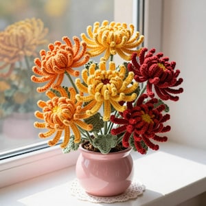 Könnte beinhalten: Ein gehäkeltes Blumenarrangement mit mehreren chrysanthemenartigen Blüten in Gelb-, Orange- und Dunkelrottönen. Die Blumen haben texturierte, gekräuselte Blütenblätter und grüne gehäkelte Blätter, präsentiert in einem glatten, hellrosa Keramiktopf.