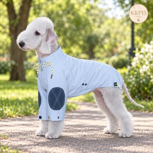 May include: A light blue dog coat featuring a high collar with yellow and white drawstring cords, dark blue circular elbow patches with cross-stitching, and silver-toned snap buttons along the front closure and hem. The text KKP15 is visible.