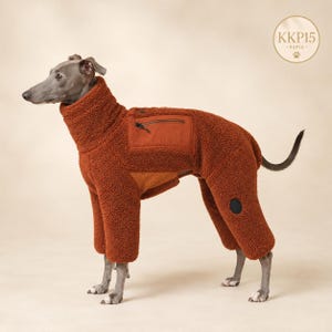 Könnte beinhalten: Ein grauer Hund trägt einen rostfarbenen Fleece-Overall mit langen Ärmeln und hohem Kragen. Das Kleidungsstück hat eine Reißverschlusstasche auf dem Rücken und einen kleinen runden schwarzen Aufnäher am Hinterbein. Ein kreisförmiges Logo mit dem Text KKP15 ist sichtbar.