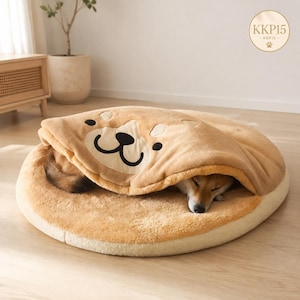 Könnte beinhalten: Ein rundes, beiges Plüsch-Haustierbett mit einer weichen, integrierten Decke, die ein Shiba-Inu-Gesicht als Cartoon-Design zeigt. Die Decke ist teilweise angehoben und gibt den Blick auf einen darin ruhenden Hund frei. Logo mit Text KKP15.
