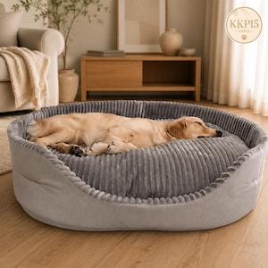 May include: A large, oval-shaped pet bed with a textured, ribbed grey fabric surface and smooth grey sides. A light-colored dog is curled up asleep in the center of the bed, which features a raised, cushioned rim for support.