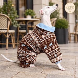 Op de afbeelding: Een witte hond draagt een bruin en crème fleece jumpsuit met een lichtblauwe kraag. De jumpsuit heeft een herhalend patroon van kleine blauwe herten, hartjes en bloemmotieven. De hond staat op een stenen ondergrond met een rond logo in de hoek.