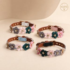 May include: Four pet collars with brown, light blue, and black leather-like straps, each decorated with a row of fabric flowers in shades of pink, grey, and dark green, featuring central clear rhinestones and gold-tone metal buckles. Text KKP15.
