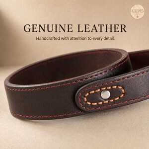 May include: A dark brown leather strap featuring red stitching along the edges and a silver-toned metal rivet. The text GENUINE LEATHER and Handcrafted with attention to every detail. are visible above the strap, along with a small circular logo.