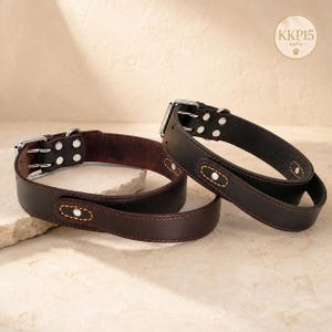 May include: Two dark brown leather collars featuring silver-toned metal buckles, rivets, and contrast stitching. Each collar has a reinforced loop detail with a central rivet. A circular logo in the top right corner displays the text KKP15.