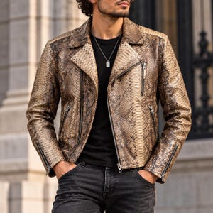 Puede incluir: Una chaqueta de motorista de cuero con estampado de piel de serpiente en tonos beige y marrón, con cuello de solapa ancho, cremalleras metálicas plateadas en la parte delantera y los puños, sobre una camiseta negra con colgante de placa.