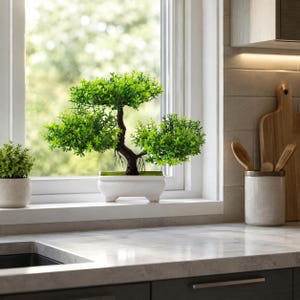 Puede incluir: Un pequeño bonsái artificial con hojas de color verde vibrante y un tronco marrón oscuro, plantado en un recipiente rectangular de cerámica blanca con cuatro patas cortas, colocado en el alféizar de una ventana junto a una planta verde más pequeña.