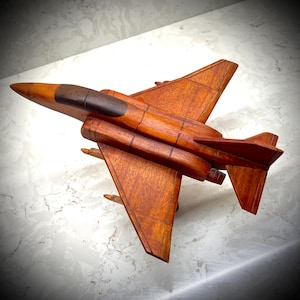 May include: A handcrafted wooden model airplane featuring a polished reddish-brown finish with a darker wood cockpit canopy. The aircraft design includes swept-back wings, a vertical stabilizer, and small under-wing attachments in a smooth wood grain.
