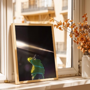 May include: A rectangular gold-toned frame containing an anime-style illustration of a figure in a green jacket looking up at a bright horizontal light beam against a dark, starry background. The artwork is displayed in a vertical orientation.