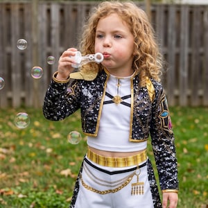 May include: A two-piece outfit consisting of a white crop top and white shorts with a gold chain print, paired with a black sequined cropped jacket featuring gold trim, shoulder accents, and colorful geometric embroidery on the sleeves.