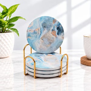 May include: A set of four circular coasters featuring a blue, white, and gold marbled pattern, stacked vertically within a gold-toned metal wire holder. The coasters have a smooth, flat surface and are arranged in a neat, upright display.