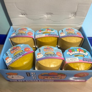 Könnte beinhalten: Ein Pappaufsteller mit sechs gelben, runden, korbartigen Behältern mit der Aufschrift Squishy Steam Bunz. Jeder Behälter ist mit einem bunten Aufkleber versehen, der den Text What Color Will Get? Squishy Fidget Fun! und One Set Sugar Edition trägt.