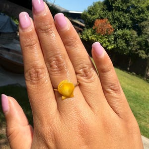 May include: A gold-toned ring featuring a small, vibrant yellow scallop shell as the central focal point. The shell has a fan-like shape with distinct vertical ridges radiating from the base, securely attached to a thin, polished metal band.