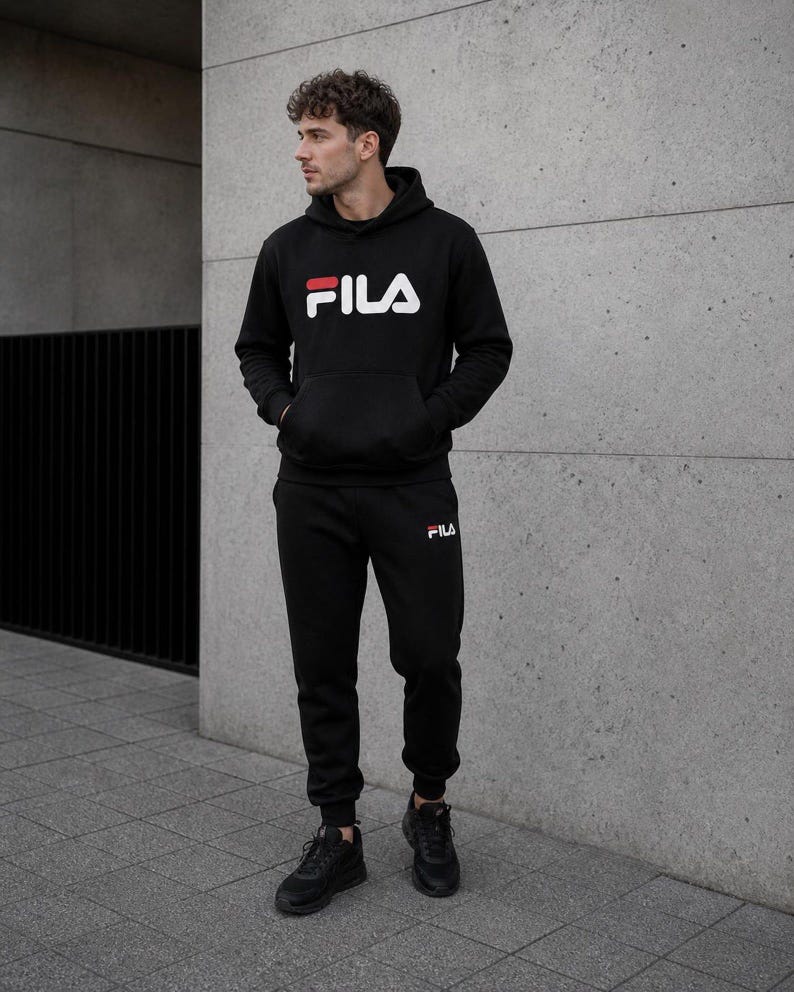 May include: A black hooded sweatshirt with a large white and red FILA logo on the chest, paired with matching black sweatpants featuring a small white and red FILA logo on the left thigh and black athletic sneakers with thick soles.