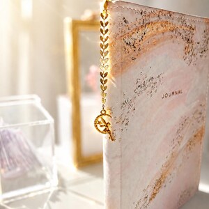 May include: A journal with a pale pink and white marble-patterned cover featuring gold foil accents and the word JOURNAL printed on the front. A gold metal bookmark with a leaf-shaped chain and a crescent moon charm with a fairy figure is attached.