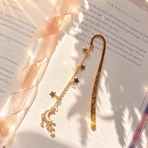 May include: A gold-toned metal bookmark featuring a flat, engraved blade and a dangling chain attachment. The chain is adorned with several small gold stars and a crescent moon charm embellished with clear, sparkling crystals at the end.