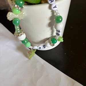 May include: A beaded bracelet featuring a mix of green, white, and clear beads, including silver-toned spacers and a light green charm. The bracelet includes white letter beads spelling out the phrase GLORY TO GOD in a circular arrangement.