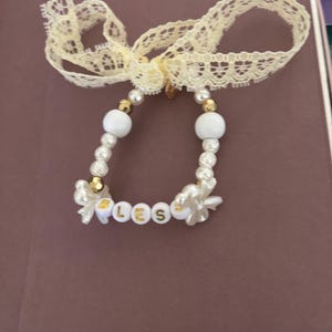 May include: A beaded bracelet featuring white and gold-toned beads, small white flower-shaped charms, and white circular beads with the gold-colored letters B, L, E, S, S printed on them. The bracelet is tied with a cream-colored lace ribbon bow.