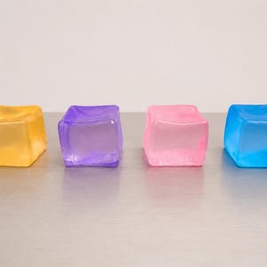 May include: Four translucent, square-shaped blocks arranged in a horizontal row. The blocks are coloured yellow, purple, pink, and blue, respectively. Each block has a smooth, slightly rounded surface and a semi-transparent, jelly-like appearance.