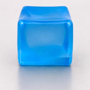 Op de afbeelding: Een klein, vierkant object gemaakt van doorschijnend blauw materiaal. Het item heeft een glad, licht concaaf bovenoppervlak met afgeronde randen en hoeken, en vertoont een uniforme, levendige azuurblauwe kleur in zijn solide, blokvorm.