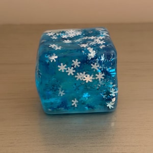 Op de afbeelding: Een doorschijnende blauwe kubus gemaakt van hars, met talloze ingebedde witte en metallic blauwe sneeuwvlokvormige glitterstukjes door de hele structuur. Het object heeft een gladde, glanzende afwerking en een vierkante vorm met afgeronde randen.