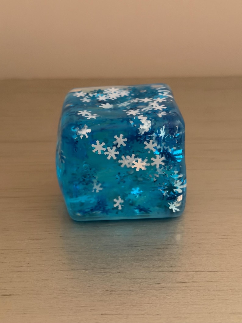 Op de afbeelding: Een doorschijnende blauwe kubus gemaakt van hars, met talloze ingebedde witte en metallic blauwe sneeuwvlokvormige glitterstukjes door de hele structuur. Het object heeft een gladde, glanzende afwerking en een vierkante vorm met afgeronde randen.