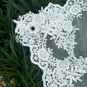 May include: A white bridal veil featuring intricate floral lace embroidery along the scalloped edges. The sheer tulle fabric is accented with a small, circular silver metal grommet integrated into the lace pattern, creating a detailed decorative border.