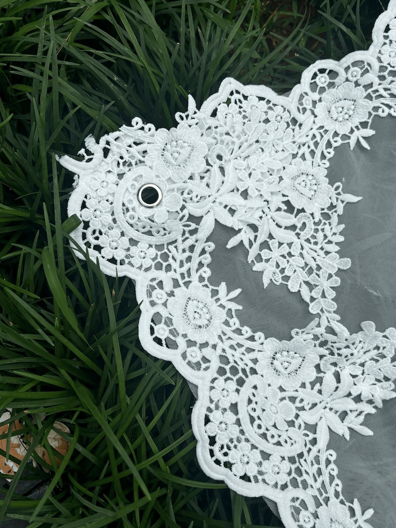 May include: A white bridal veil featuring intricate floral lace embroidery along the scalloped edges. The sheer tulle fabric is accented with a small, circular silver metal grommet integrated into the lace pattern, creating a detailed decorative border.