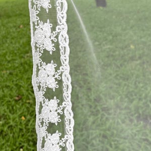 May include: A white wedding veil featuring a sheer tulle fabric and a decorative lace border. The lace trim displays a repeating floral pattern with embroidered blossoms and leaves, creating a detailed edge along the length of the translucent veil.