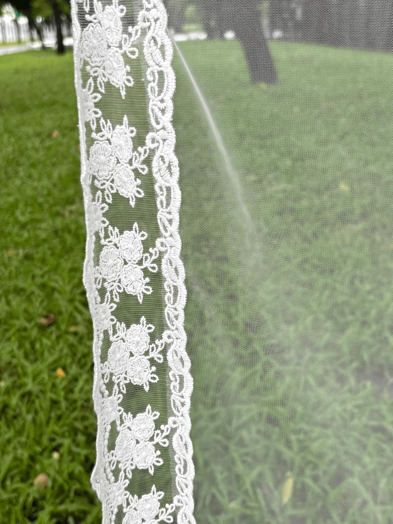 May include: A white wedding veil featuring a sheer tulle fabric and a decorative lace border. The lace trim displays a repeating floral pattern with embroidered blossoms and leaves, creating a detailed edge along the length of the translucent veil.