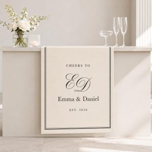 Może przedstawiać: Kremowy bieżnik na stół z czarną obwódką z podwójnej linii i wyśrodkowanym czarnym napisem CHEERS TO, monogramem ED, imionami Emma & Daniel oraz napisem EST. 2026, umieszczony na blacie ze szklanym wazonem z białymi kwiatami.