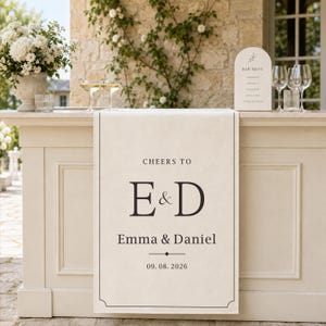 Peut inclure: Un chemin de table en tissu crème avec un texte imprimé noir indiquant CHEERS TO, E & D, Emma & Daniel, et 09.08.2026. Le chemin de table présente une fine bordure noire avec des accents décoratifs aux coins et pend devant un comptoir blanc.
