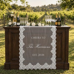 Könnte beinhalten: Ein weißer Spitzen-Tischläufer, der über eine dunkle Holzbar drapiert ist, mit dem aufgedruckten Text CHEERS TO The Harrisons EST. 2026. Der Läufer ist mit komplizierten floralen Spitzenrändern verziert und von Weinflaschen und Gläsern umgeben.