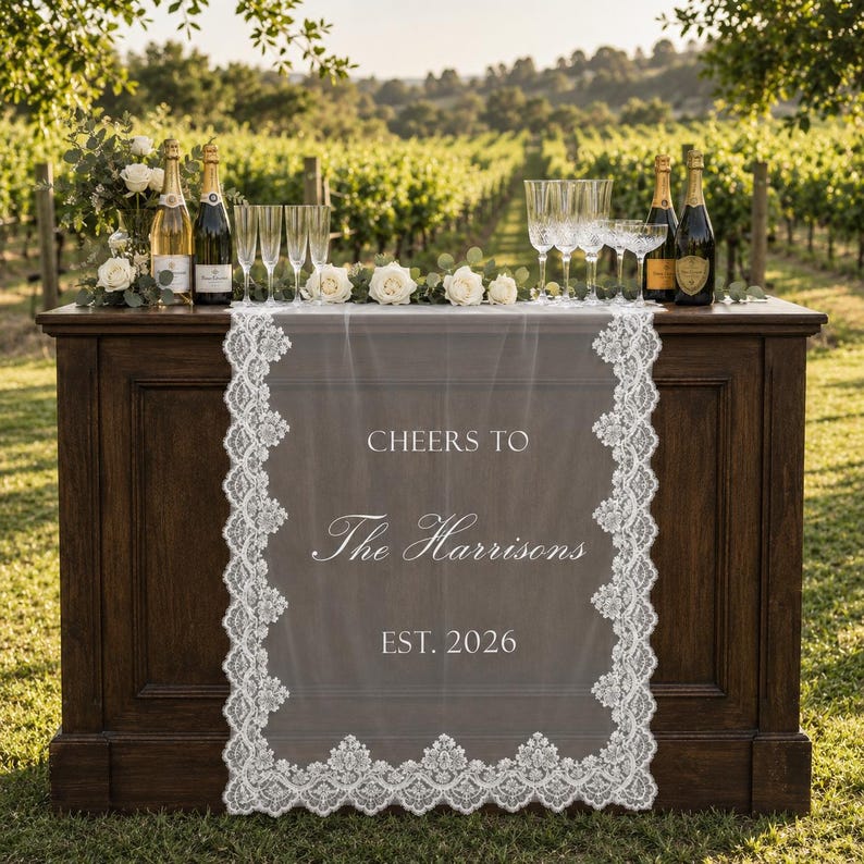 May include: A white lace table runner draped over a dark wooden bar, featuring the printed text CHEERS TO The Harrisons EST. 2026. The runner has intricate floral lace borders and is surrounded by champagne bottles, clear glasses, and white roses.