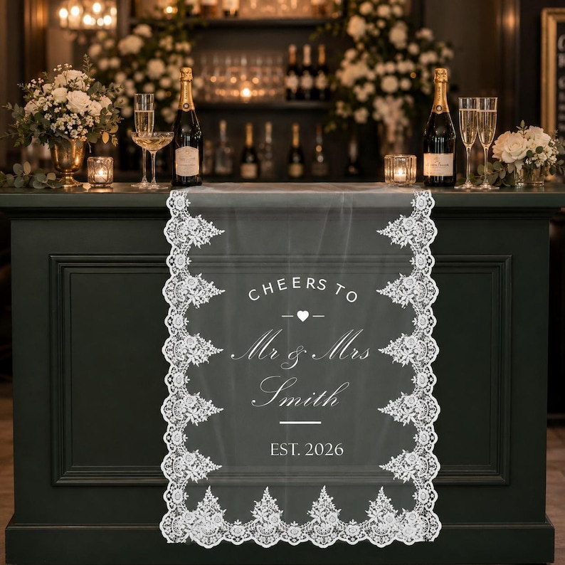 May include: A white lace table runner featuring a scalloped floral border and centered white text that reads CHEERS TO Mr & Mrs Smith EST. 2026 with a small heart icon. The runner is draped over a dark green bar counter with champagne bottles.