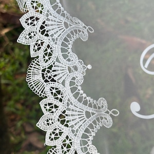 May include: A white bridal veil featuring an intricate scalloped lace edge with detailed floral and fan-like embroidery patterns. The sheer, lightweight tulle fabric extends from the decorative lace border, showcasing a delicate and fine mesh texture.